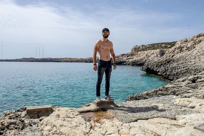 Full length of shirtless man standing on rock against sky