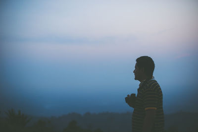 Side view of young man holding camera looking away while standing on field against sky