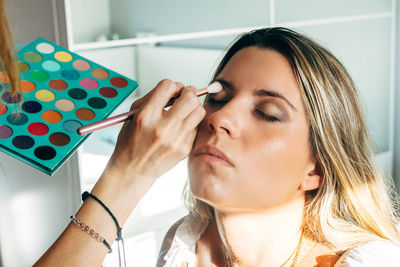 Unrecognizable female makeup artist applying stylish eyeshadows on client with eyes closed in bright beauty salon