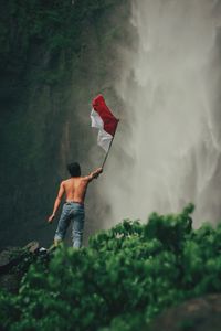 Rear view of man standing by waterfall with indonesian national flag