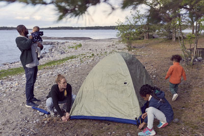 Father carrying baby boy while woman and daughter setting tent at beach