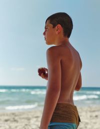 Side view of shirtless boy holding food while standing on beach against sky