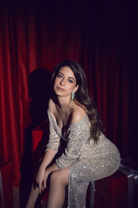 Beautiful young woman in a shiny dress is sitting on red gift boxes with bows in a spot of light