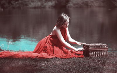 Full length of young woman in red evening gown opening box at lakeshore