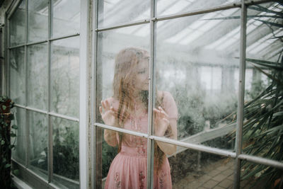 Through glass of serene young female in dress standing in hothouse with green plants