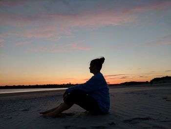 Man sitting on beach against sky during sunset