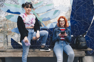 Portrait of hipster female friends holding mobile phones against mosaic wall