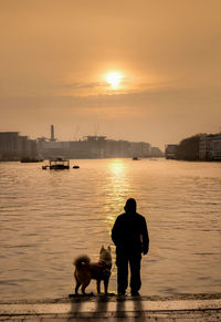 Silhouette man with dog by river against sky during sunset