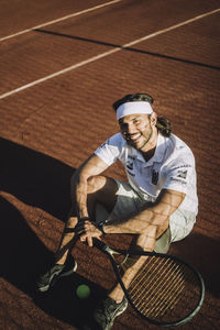 High angle view of smiling man with tennis racket sitting on ground during break