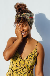 Smiling african american female wearing summer dress and trendy headband standing near wall in city and touching face while looking at camera