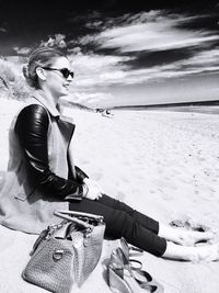 Woman wearing sunglasses sitting on sand at beach against sky