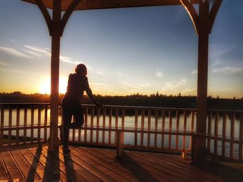 Woman standing in gazebo by river against sky during sunset