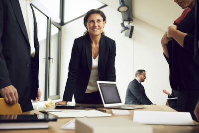 Smiling mature businesswoman leaning on conference table while standing amidst lawyers at office during meeting