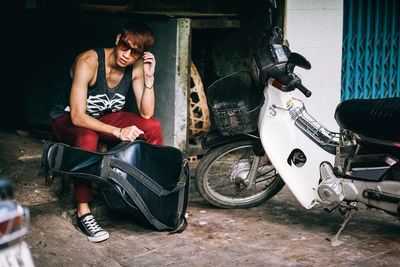 Portrait of young man sitting with guitar by motor scooter on footpath