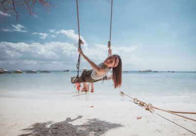 Portrait of smiling young woman sitting on swing at beach against sky