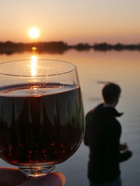 Close-up of wine holding glass against sea during sunset