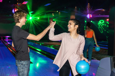 Smiling multi-ethnic friends giving high-five on illuminated parquet floor at bowling alley