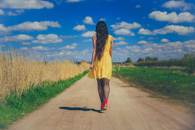 Rear view of woman walking on road amidst field against sky