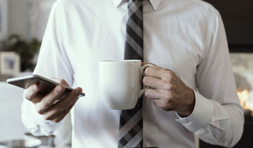 Midsection of businessman holding coffee cup using smart phone while standing in office