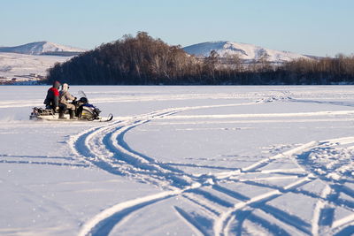 The snowmobile with fishermen is riding along the winter lake against a hill with birches.