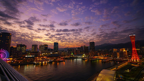 Illuminated buildings by river against sky during sunset