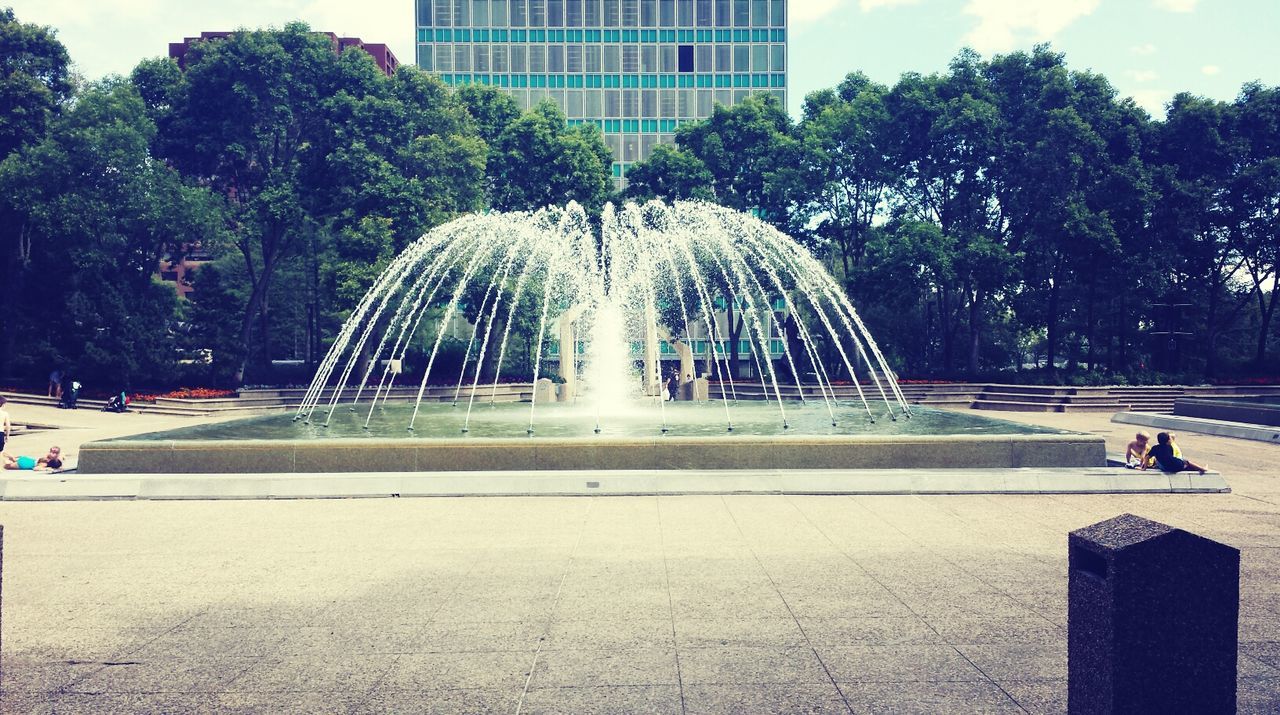 tree, fountain, architecture, built structure, building exterior, park - man made space, incidental people, city, motion, spraying, modern, outdoors, day, water, leisure activity, sculpture, growth, city life, reflection