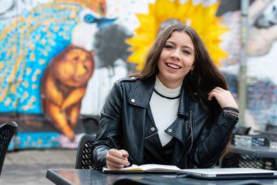 Amazing optimistic young woman with long hair taking notes in notebook while sitting in outdoor cafe