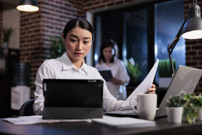 Businesswoman holding paper looking at digital tablet screen in office