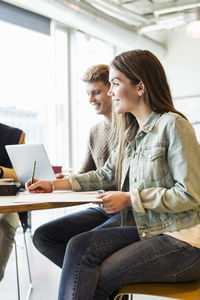 Happy woman writing while sitting with friend in university classroom