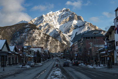 Snow covered road amidst buildings and mountains against sky