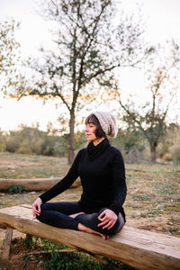 Smiling woman wearing knit hat meditating on bench at park