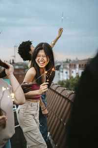 Cheerful young woman holding lit sparkler enjoying with friends on rooftop