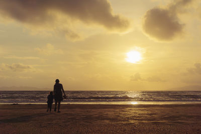 Rear view of silhouette mother and daughter walking at beach against sky during sunset