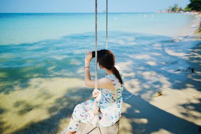 High angle view of woman photographing sea while sitting on swing at beach
