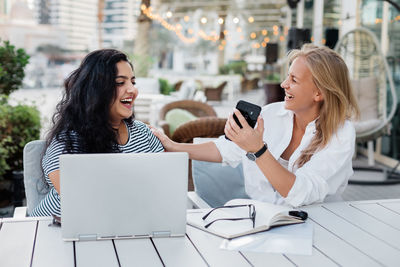 Two young women laugh during a break in a cafe, look through working materials on a laptop 