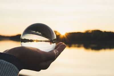 Close-up of hand holding crystal ball against sky during sunset