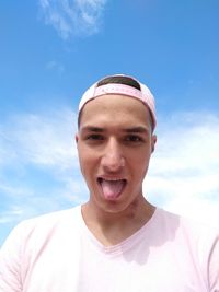 Portrait of young man wearing cap while sticking out tongue against sky
