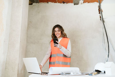 Professional confident architect woman in construction site using mobile phone holding blueprints