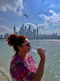 Red haired women drinking water on the beach, downtown dubai in background, bird flying over