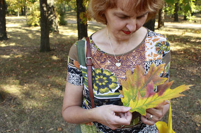 Senior woman holding maple leaves while standing on field against trees in park during autumn