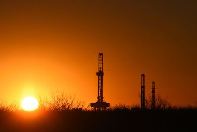 Silhouette drilling rigs on field against clear sky during sunset