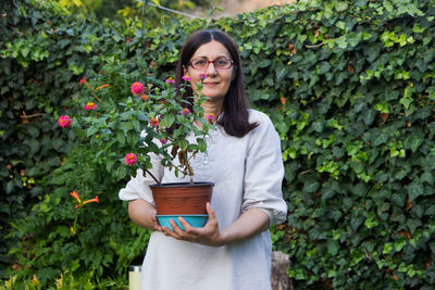 Portrait of woman holding potted plant while standing against plants