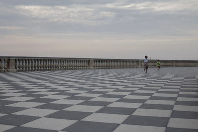 Rear view of people walking on tiled floor by sea against sky