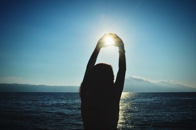 Optical illusion of woman making heart shape at beach against sky during sunset