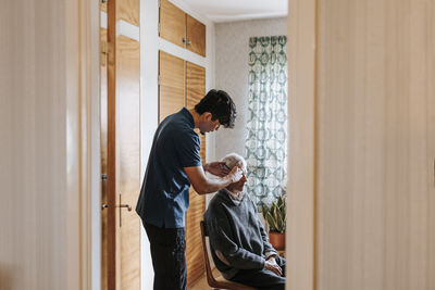 Side view of male care assistant combing hair of senior man sitting at home