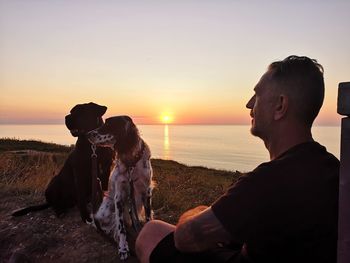 Man with dogs sitting on land against sky during sunset