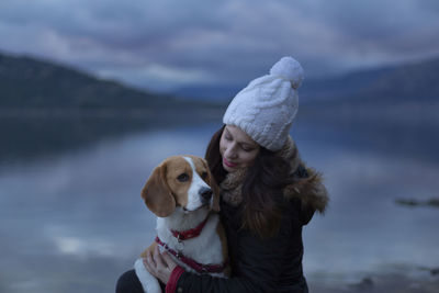 Smiling woman embracing beagle sitting by lake against sky during winter