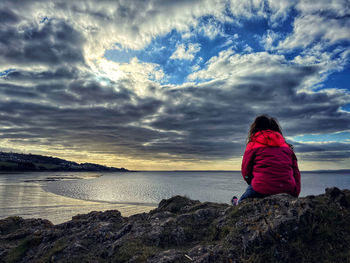 Rear view of woman sitting on rock by sea against sky