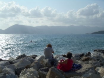 Rear view of women sitting on rock by sea against sky