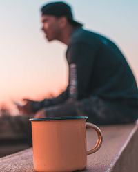 Close-up of coffee cup with man sitting in background during sunset